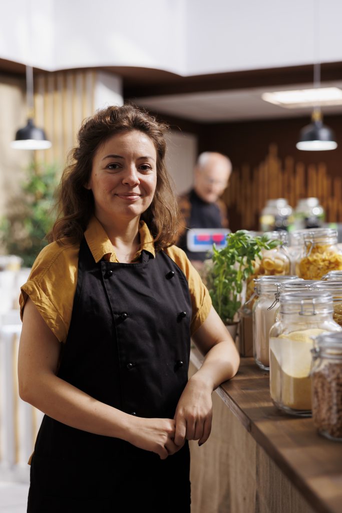 Portrait of smiling zero waste shop storekeeper next to ethically sourced low carbon footprint bulk products. Joyful woman working in green living local neighborhood store using renewable energy